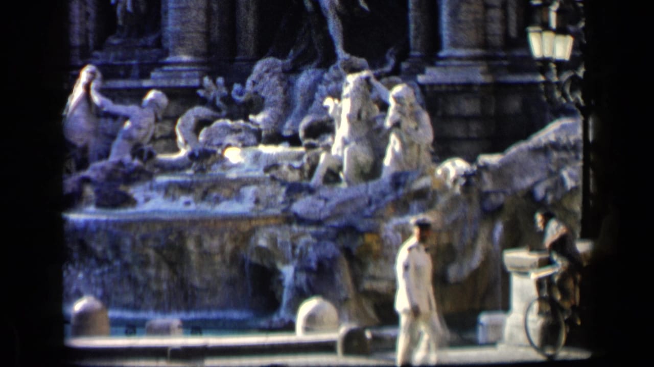 Tourists pass in front of the Trevi Fountain with cascading water and stone figures behind them.
