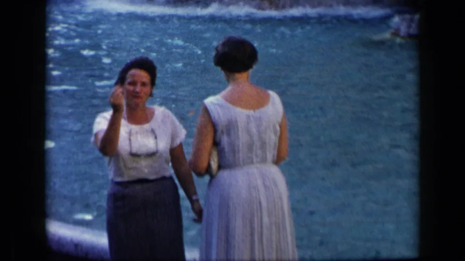 Two women stand near the fountain water in circa-1960 Italy; one in a white blouse faces the camera while the other wears a light dress.