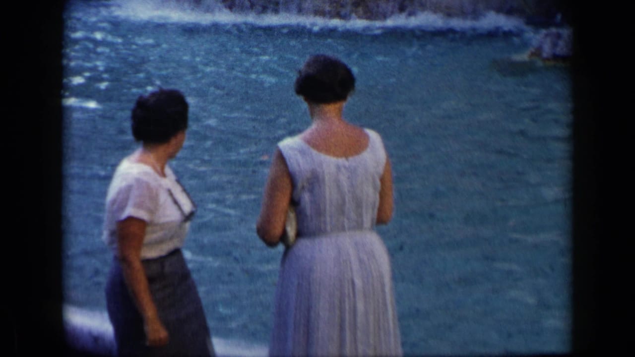 Two adult women stand near rippling water, one in a white top and dark skirt and one in a light sleeveless dress.