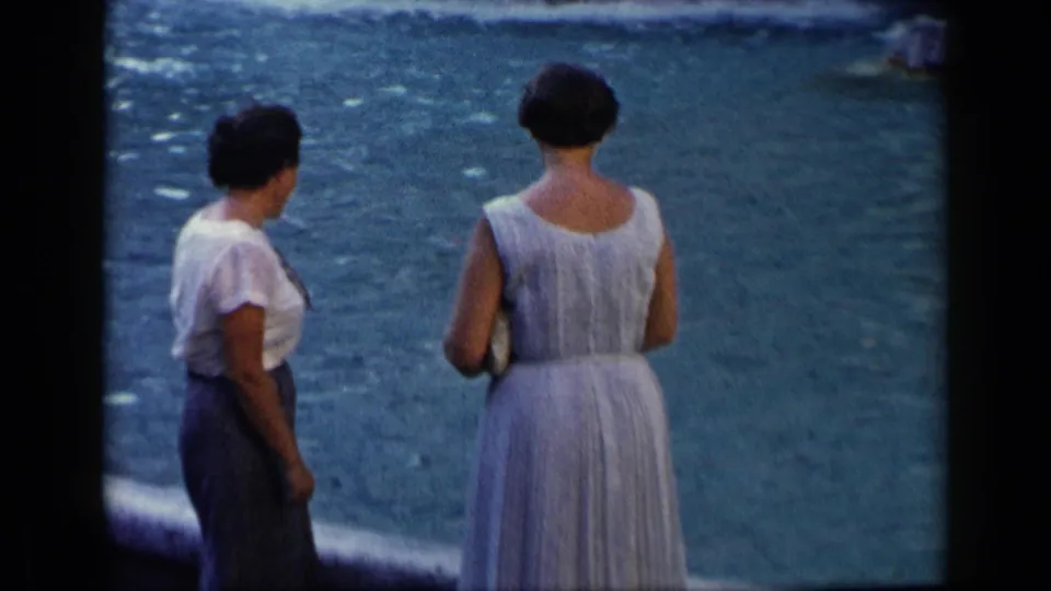 Two adult women in summer clothing stand by blue water in a 1960 Vatican, Italy home-movie frame with soft focus and grain.