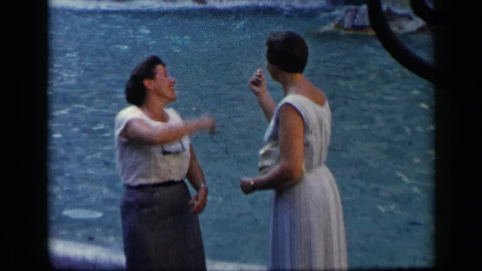 A 1960 Vatican, Italy home-movie frame captures two adult women gesturing and laughing beside a body of water.