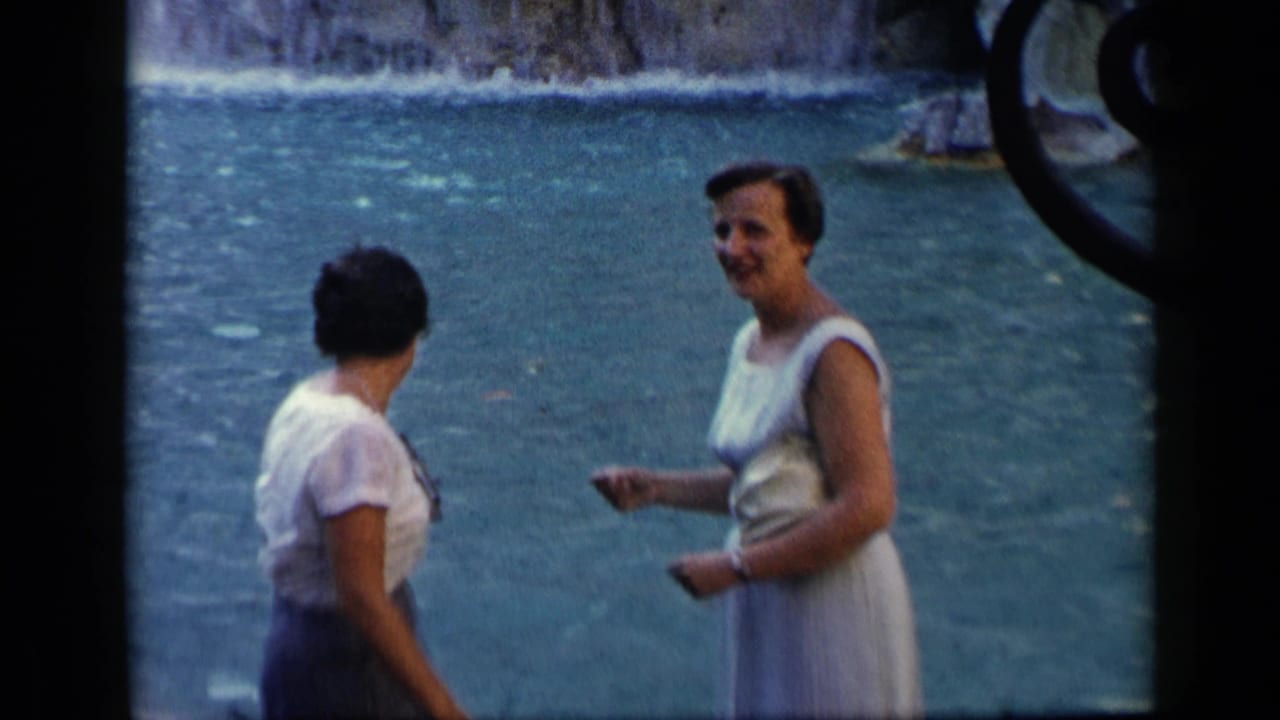 Two adult women stand beside blue-green water, one facing away and one in a light dress smiling.