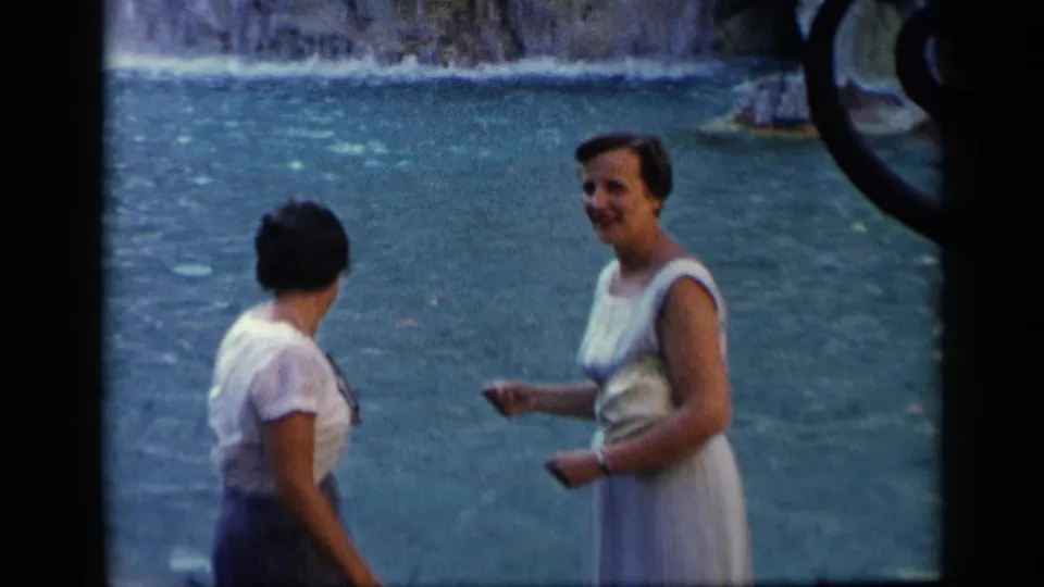 1960 Vatican, Italy home-movie frame showing two adult women conversing beside blue-green water near a stone edge.