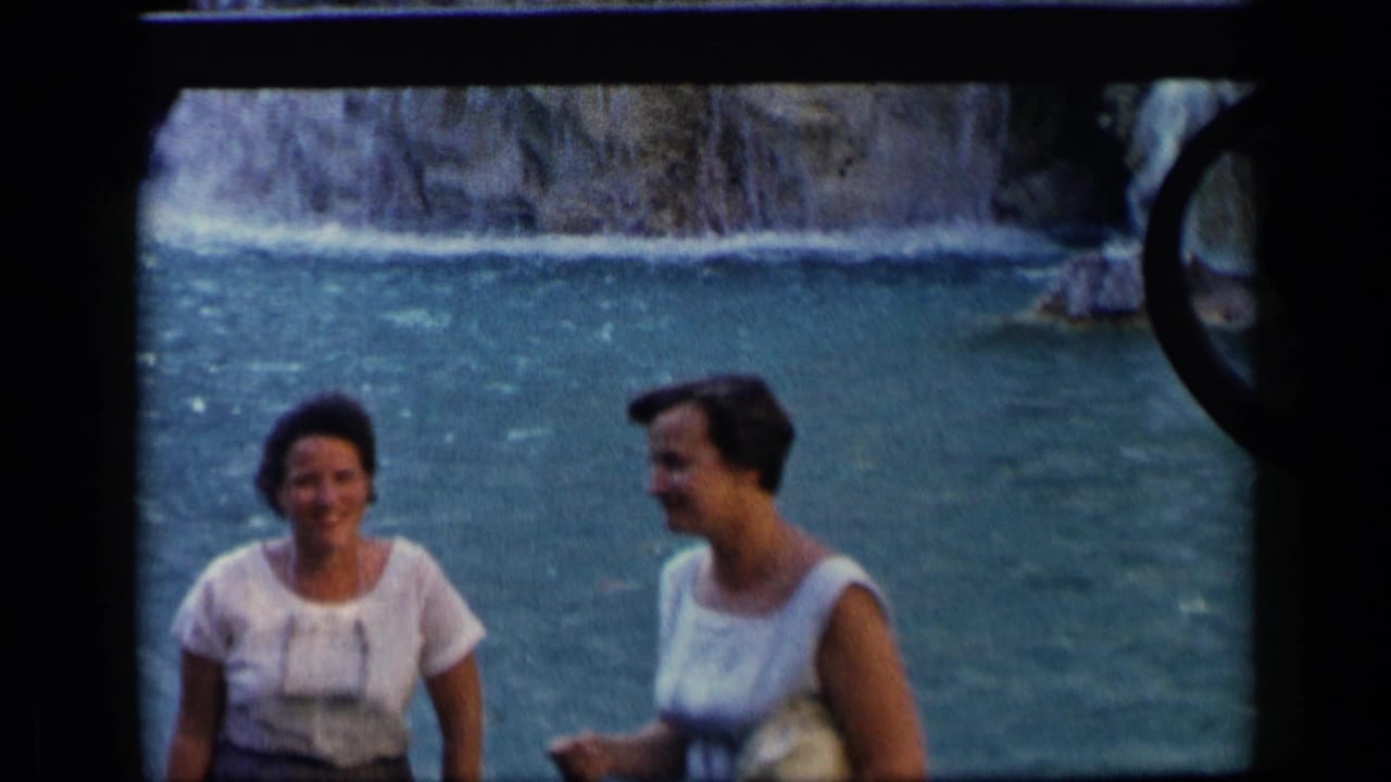 Two adult women stand near water, with one woman facing the camera and another in profile.