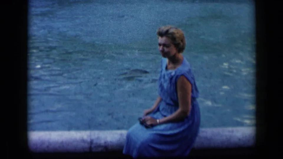 1960 Vatican, Italy home-movie frame showing an adult woman in a blue dress seated alone at the water's edge.