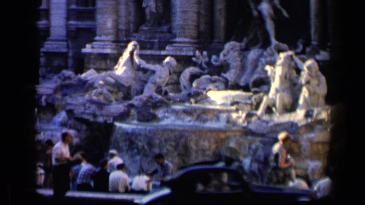 Tourists gather beside a sculpted fountain while a dark car passes in the foreground.