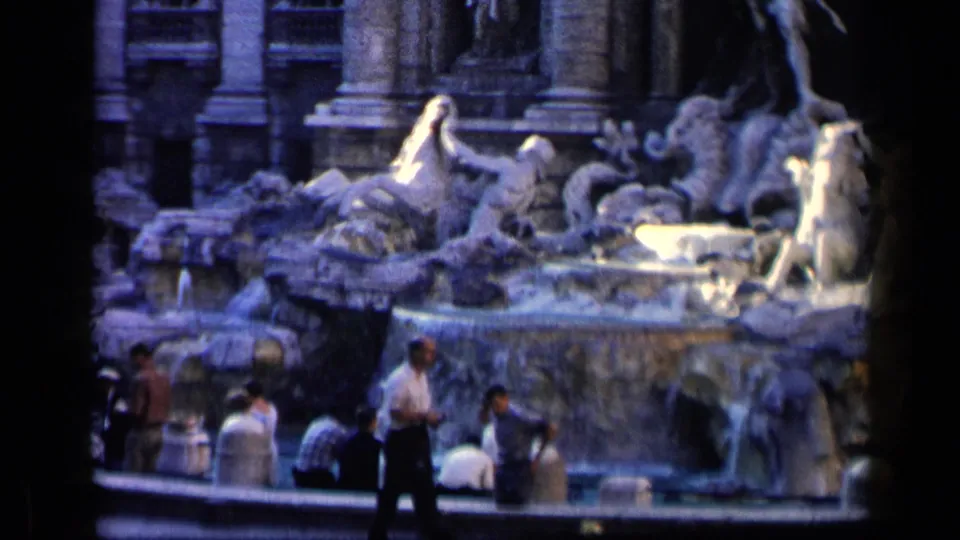 1960 8mm frame of a crowded Italian fountain scene, with a standing man, seated visitors, sculpted stone figures, and water in the basin.