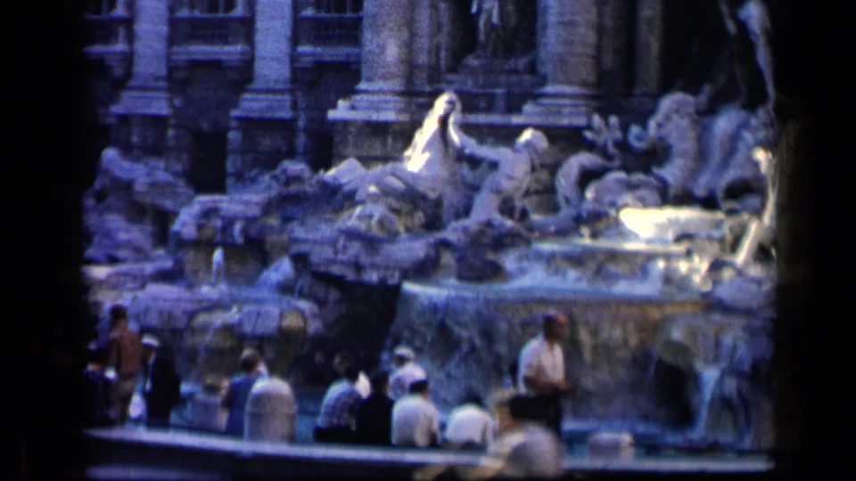 1960 8mm travel frame showing a busy fountain with seated tourists, sculpted figures, flowing water, and soft handheld framing.