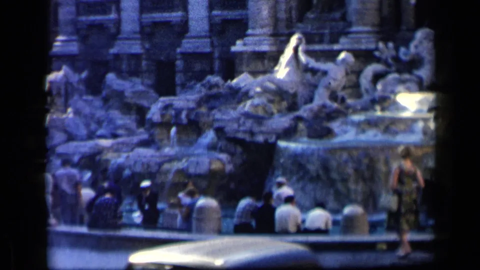 1960 8mm frame of visitors seated and standing along a monumental Italian fountain, with sculpted figures and a vehicle edge in the foreground.
