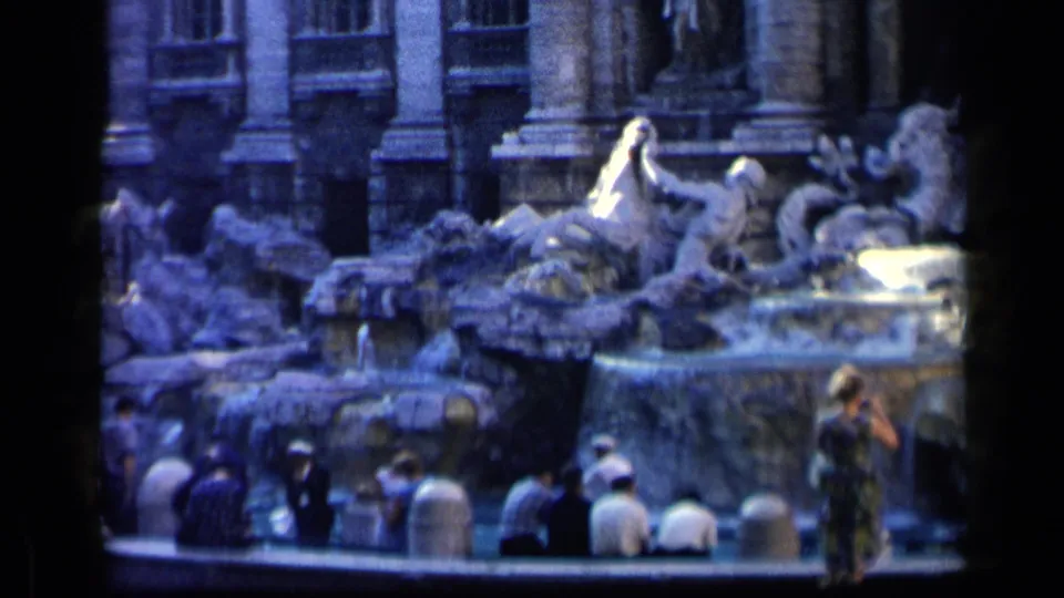 Tourists in mid-century clothing pause at the Trevi Fountain in Rome, some seated along the edge and one standing near the water.