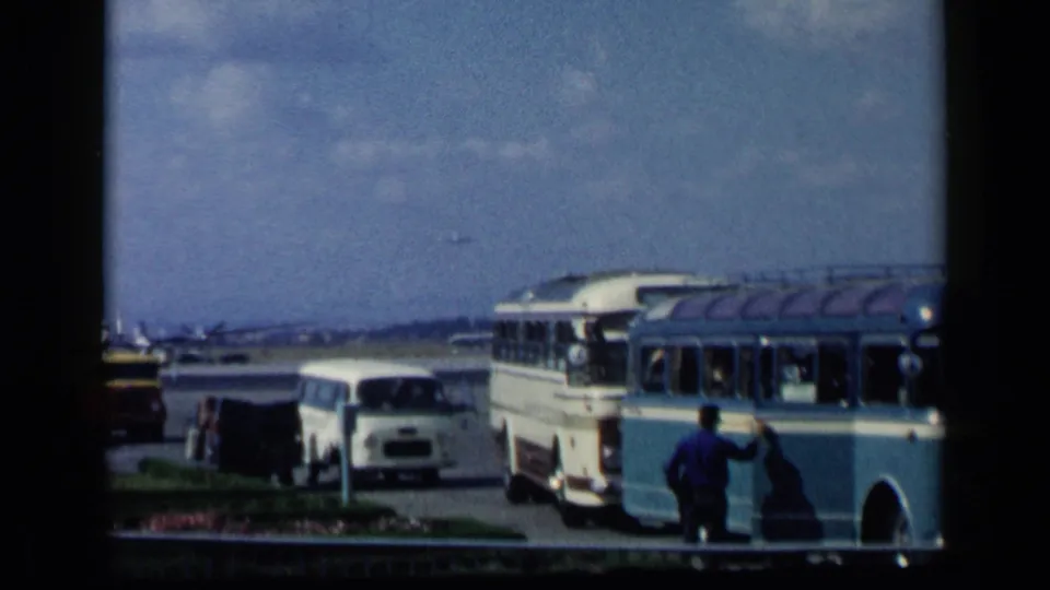 A soft 1960 8mm frame shows airport ground transport in Italy, including a blue and white bus and a man near the curb.