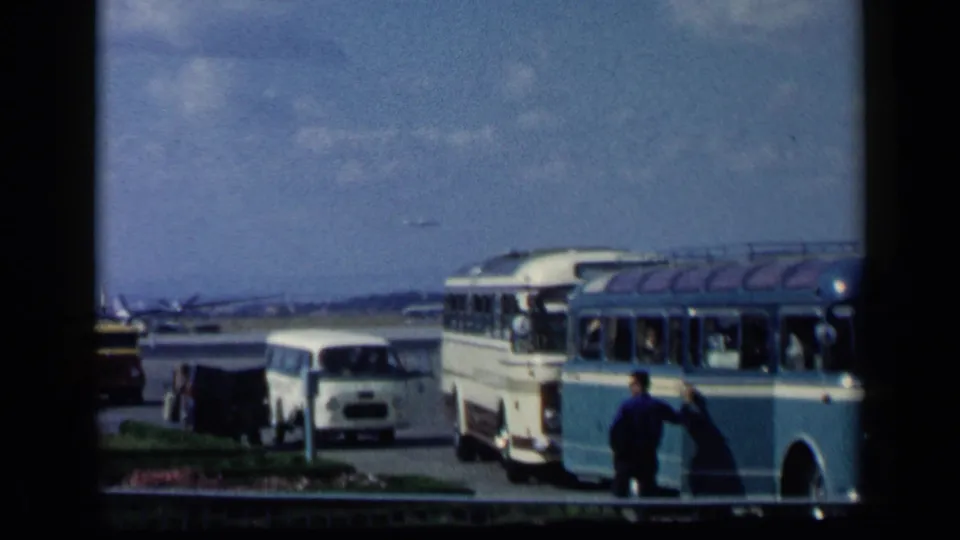 1960 Italy home-movie frame of a man beside an airport bus, with a white van, distant aircraft, pavement, and sky visible.