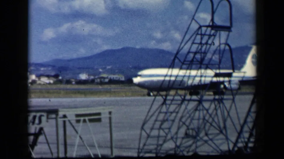 A 1960 airport tarmac frame shows the Pan Am jet partly obscured by tall mobile stairs, with hills and runway structures beyond.