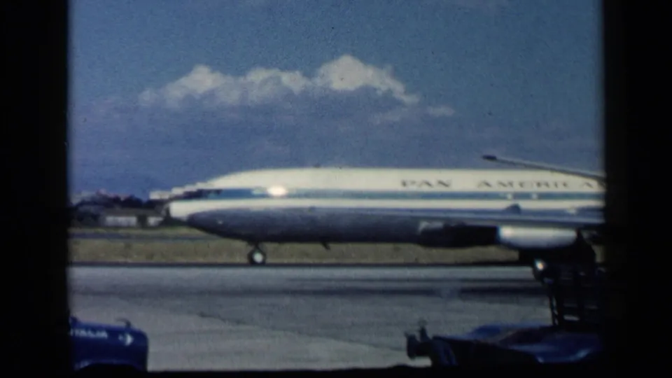 Metadata-supported 1960 airport footage shows the side of a Pan American Boeing 707 taxiing past runway equipment under clouds.