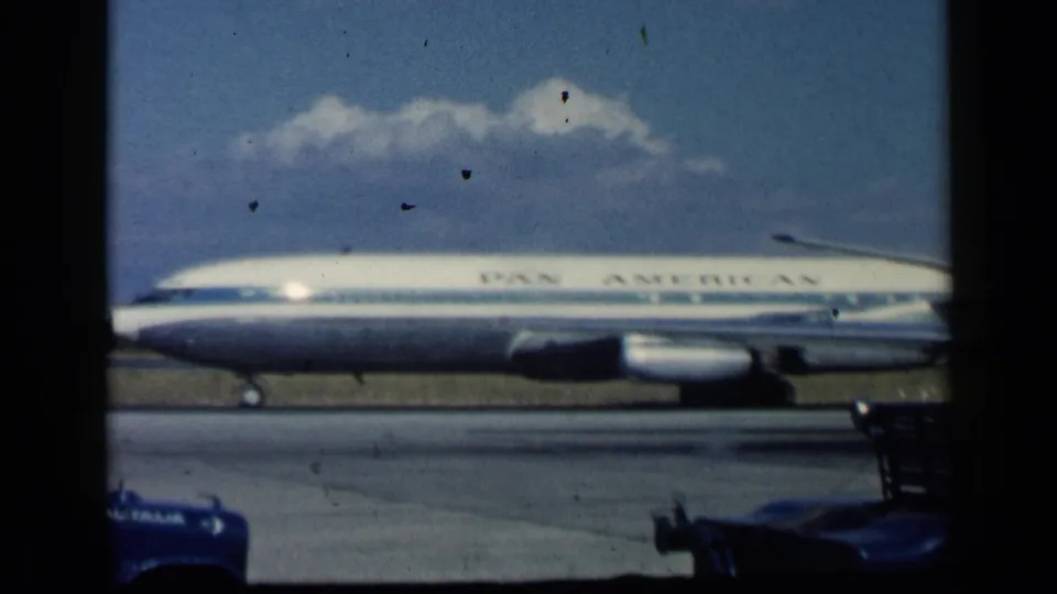 1960 archival 8mm frame showing a Pan American airliner on a runway, seen through dark film-frame borders.