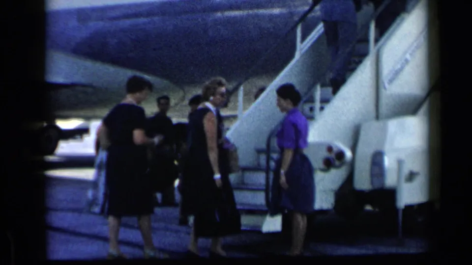 1960 archival 8mm frame showing adults, including women in dresses, near boarding stairs beside a Pan American plane.