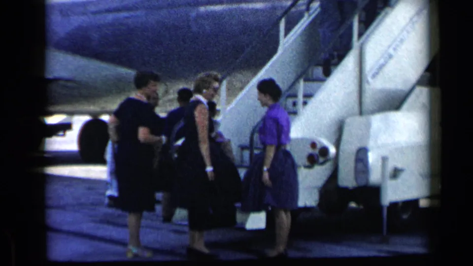 In 1960 Vatican, Italy metadata context, women in dresses carry luggage near a commercial airplane stairway on the tarmac.