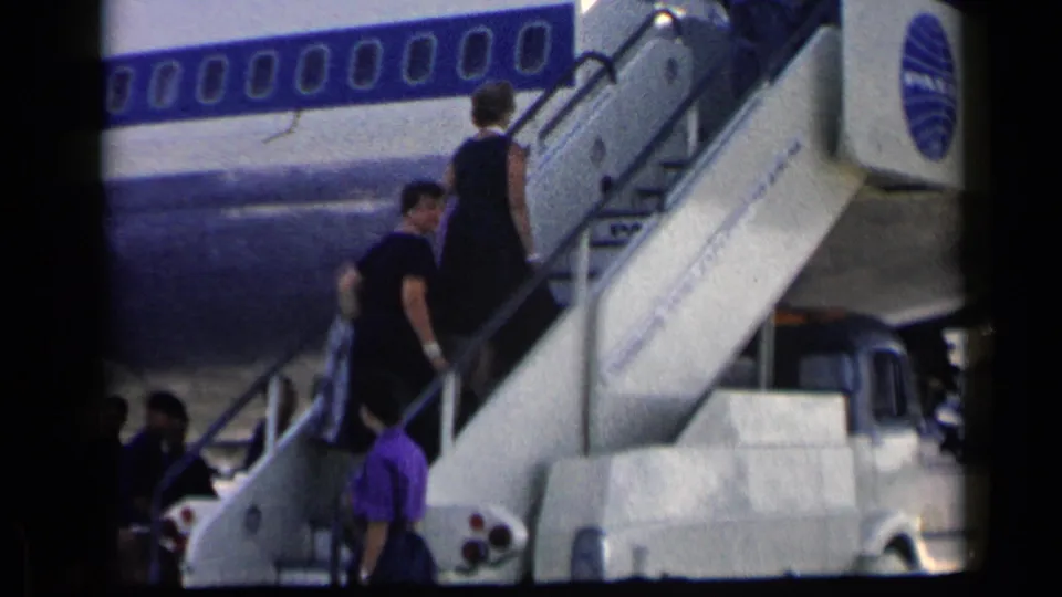 A woman in a dark dress ascends the mobile stairs to a blue-and-white aircraft as another person stands below.