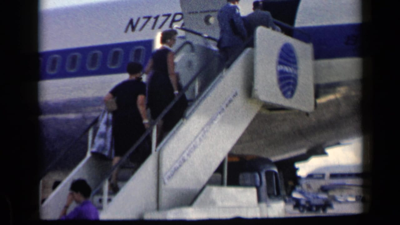 Passengers climb mobile stairs toward a Pan Am aircraft door beside a white fuselage with a blue stripe.