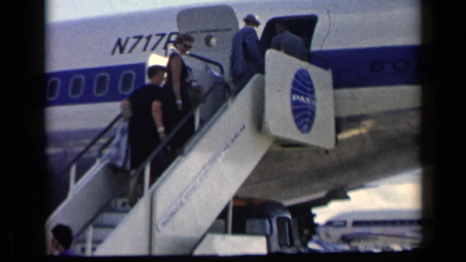 Travelers in mid-century clothing climb the mobile stairs into the Pan Am aircraft, with the globe logo and open door in view.