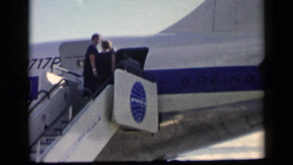 The 1960 home-movie frame shows passengers near the top of mobile stairs entering a Pan Am Boeing aircraft on the tarmac.