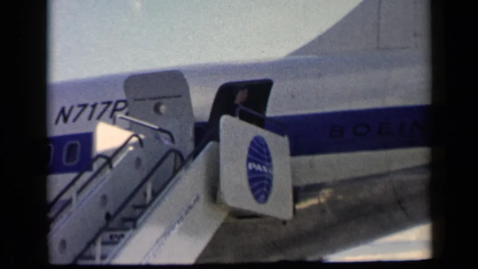 1960 8mm frame showing a Pan Am-marked Boeing with open door, boarding stairs, and partial registration visible at an airport.