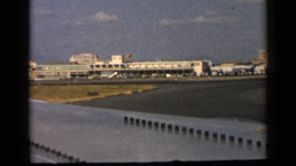 Airport terminal buildings and tarmac are seen past a partial aircraft wing in this 1960 Italy travel footage frame.