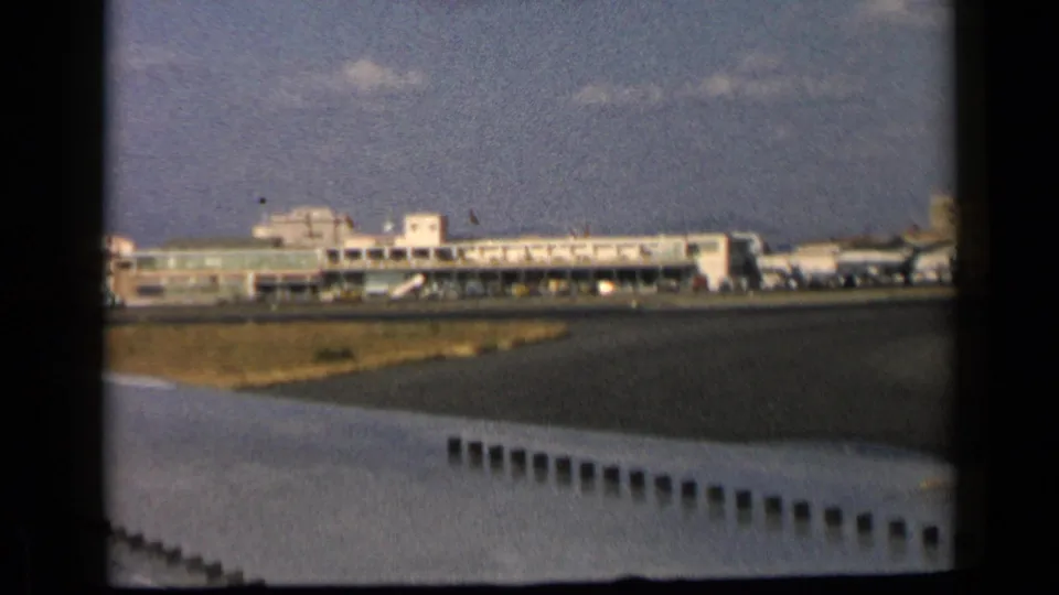 1960 8mm frame showing airport terminal structures, runway edge, grass, and an aircraft wing from a passenger-side perspective.