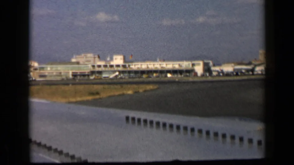 A terminal and parked aircraft are visible beyond a foreground wing in a 1960 Italy airport home-movie frame.