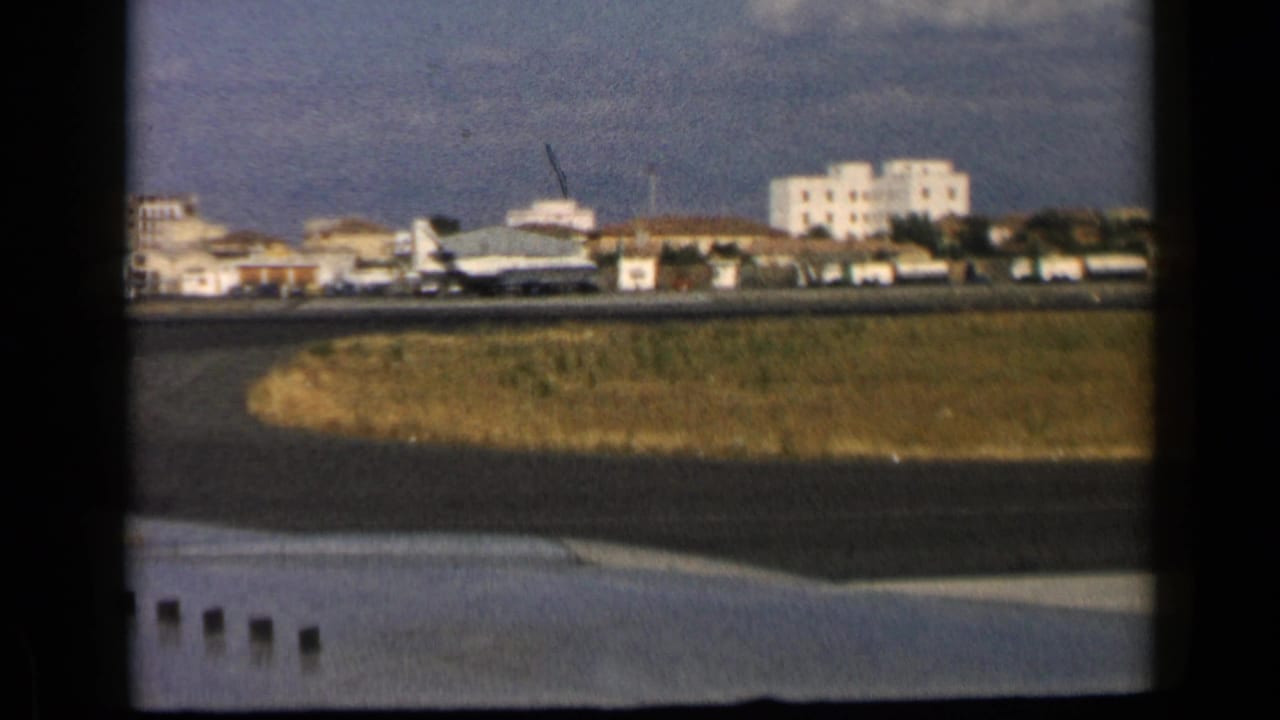 Commercial jet on a runway with grass, airport buildings, and pale residential blocks beyond.