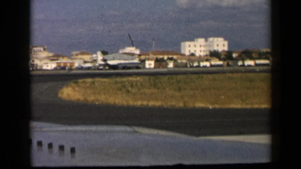 1960 8mm frame showing a commercial jet near an airport runway in the reported Vatican, Italy travel footage, with buildings in the distance.