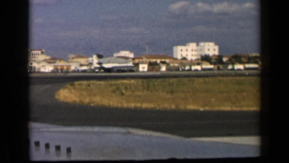 1960 8mm airport view from the reported Vatican, Italy clip, with a jet on the tarmac and pale buildings beyond the runway.