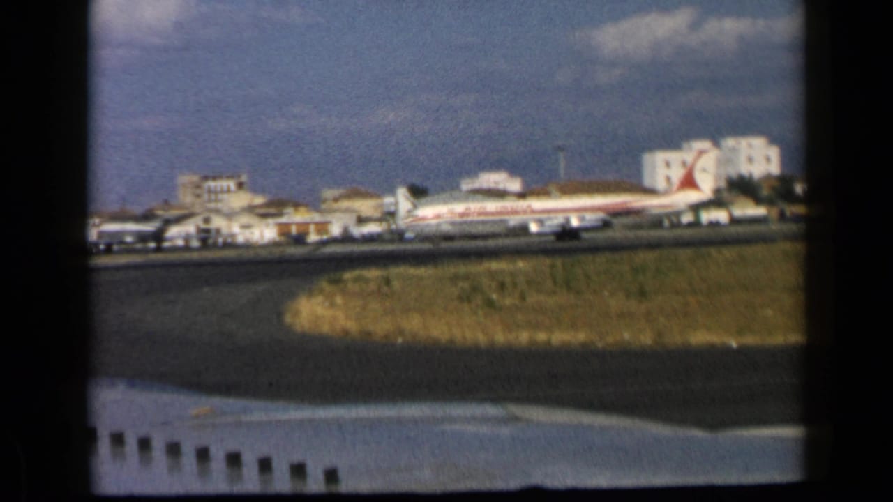 Passenger plane parked near airport buildings beyond runway grass and dark window framing.