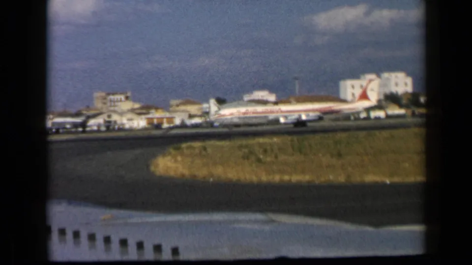 1960 home-movie view of a red-and-white passenger plane near low airport buildings, seen across runway grass through dark window framing.