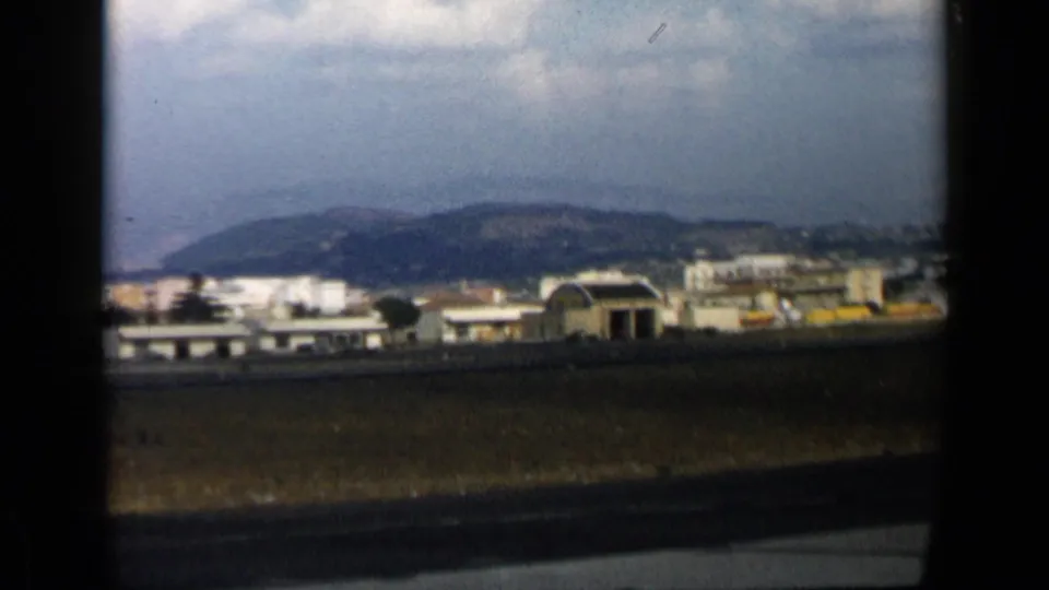 Airport grounds in Italy, with low buildings, runway pavement, grassy foreground, distant hills, and dark aircraft-window framing.