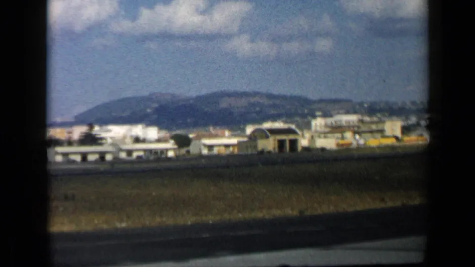 Home-movie frame showing low airport buildings, a hangar-like structure, runway pavement, grass, and hills beyond the airfield.