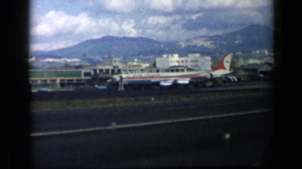 1960 airport view with a red-and-white passenger plane near terminal buildings, runway pavement in front, and mountains under a partly cloudy sky.
