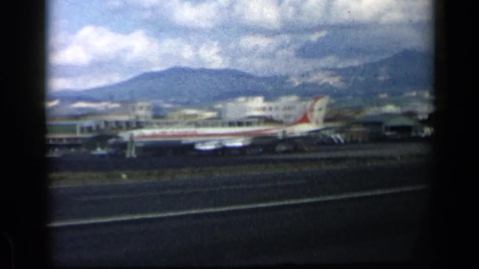 1960 8mm frame of a commercial airplane near a runway in Italy, seen through dark window edges with mountains and clouds beyond.