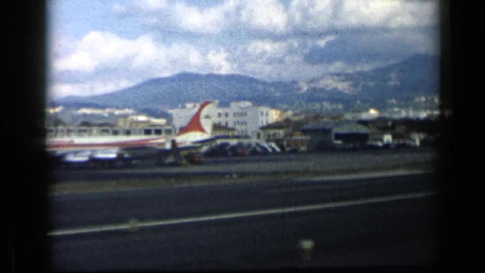 Vintage 8mm view of a red-tailed commercial aircraft by airport buildings, with a mountain ridge and partly cloudy sky behind.