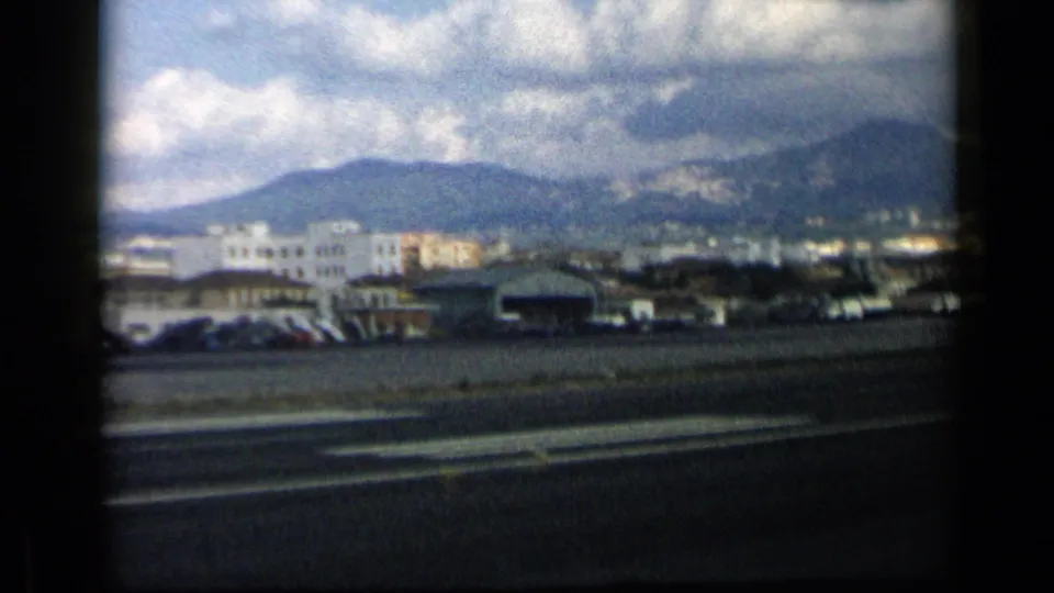 1960 home-movie frame showing airport structures and parked vehicles beyond the runway, with mountains partly obscured by clouds.