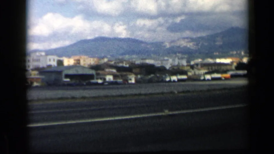 End frame of the 1960 Italy airport view, showing runway pavement, low buildings, parked vehicles, and a mountain backdrop.