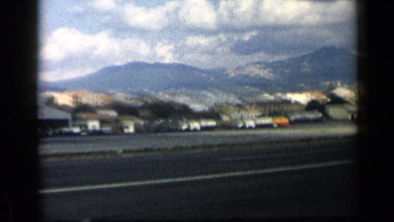 Roadway with cars, low buildings, distant hills, and cloudy sky seen through 8mm frame edges.
