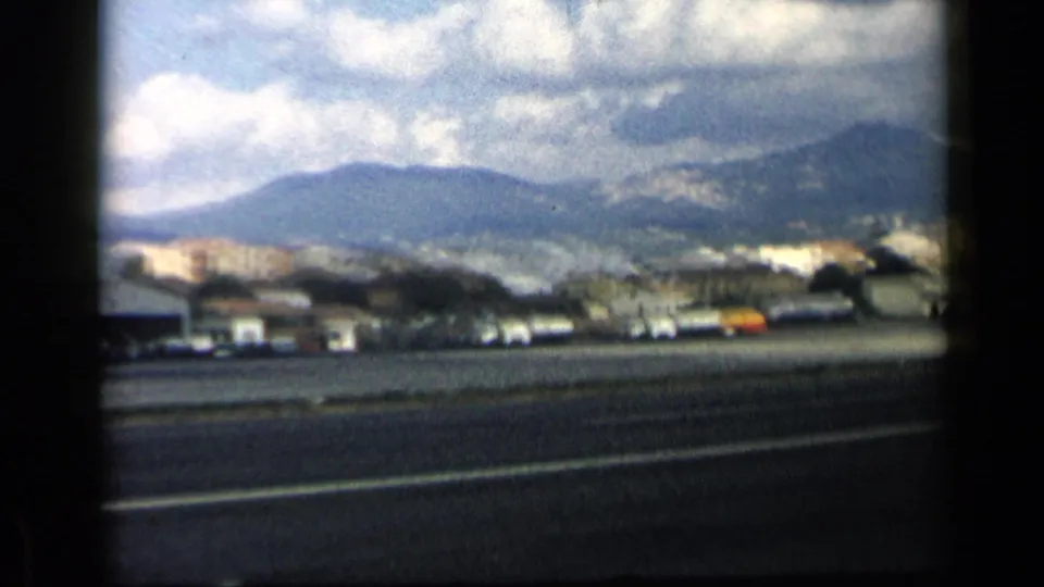 1960 8mm travel frame near Vatican, Italy metadata showing roadside traffic, low buildings, distant hills, and cloudy sky.