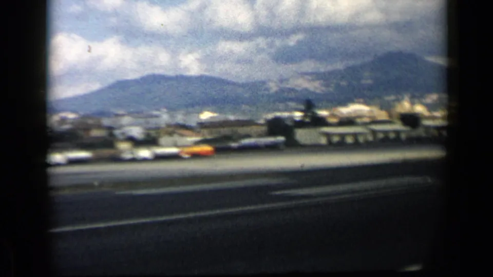 1960 8mm frame near Vatican, Italy metadata with cars along a broad road, low structures, distant hills, and heavy clouds.