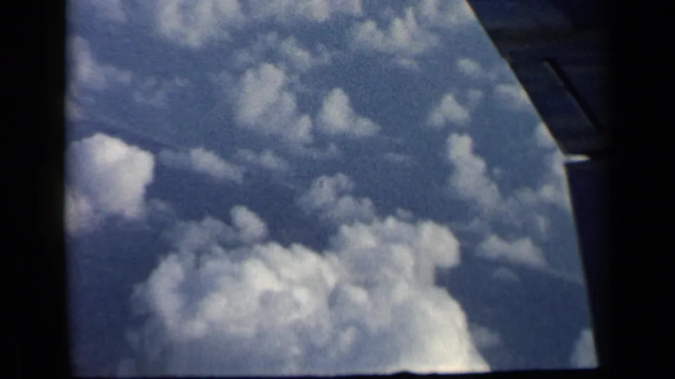 1960 8mm travel frame looking from an airplane window toward the wing edge, blue sky, and white clouds below.