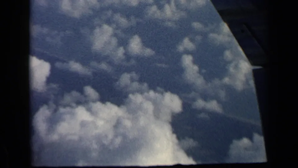 1960 8mm airplane-window view showing cloud fields, blue sky, and a partial wing or window structure at the right edge.