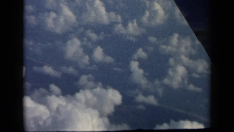 1960 8mm view of blue sky and white cumulus clouds through an airplane window, with dark film and cabin edges visible.