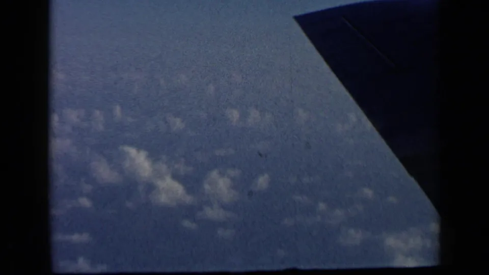 1960 8mm aerial view with an aircraft wing cutting across pale blue sky and scattered clouds below.