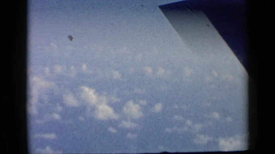 1960 8mm airplane-window view with a wingtip at the upper right and small clouds across a hazy blue sky.