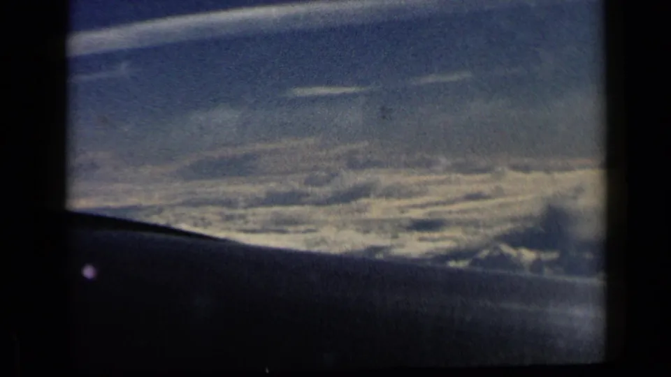 1960 8mm aerial view through an aircraft window, showing layered clouds, pale horizon, and a dark wing or window edge below.